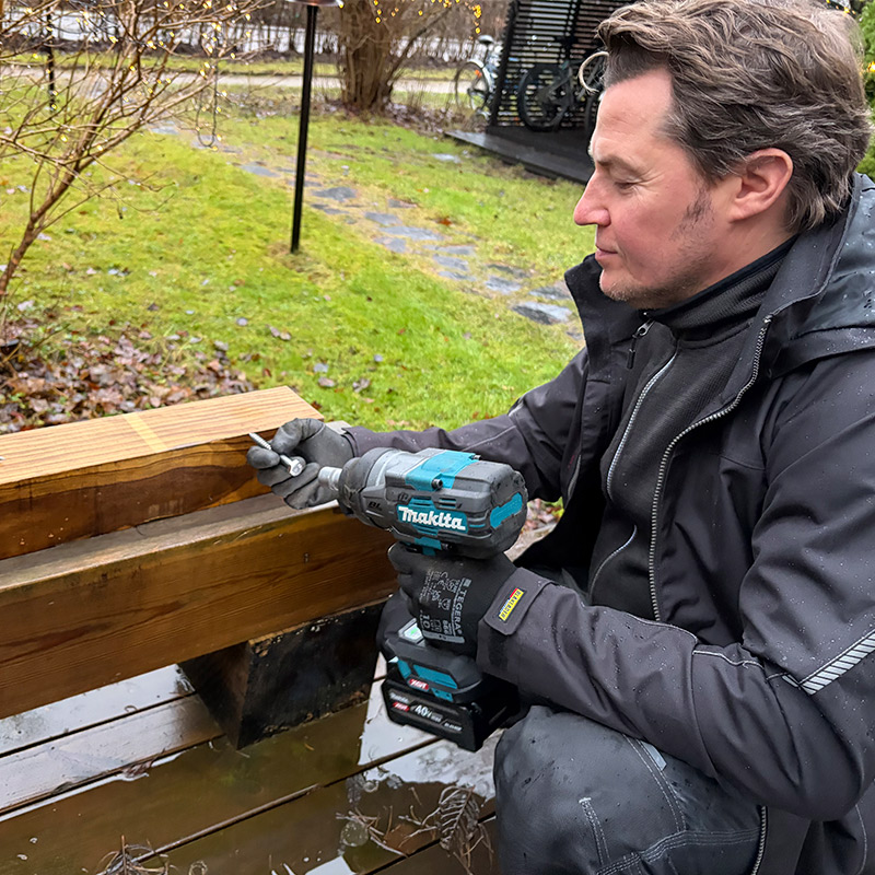 Henkilö kiinnittää pitkää ruuvia puiseen rakenteeseen Makita TW002GZ 40V XGT -mutterinvääntimellä ulkona.