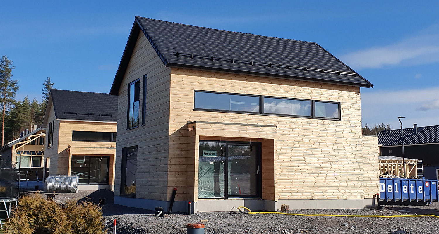 Kaksi tummakattoista vaaleaa puutaloa rakennustyömaalla, taustalla sinitaivas ja metsä.