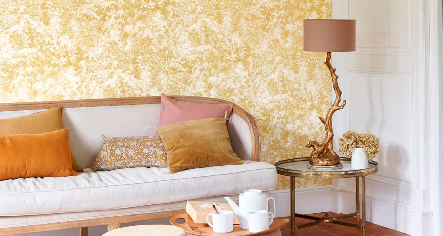 Vaalea sohva oranssi- ja roosatyynyin keltaisella tekstuuriseinällä, kultainen sivupöytä ja lamppu