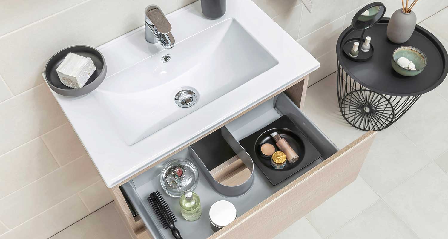 Valkoinen allas ja avonainen laatikko harmaine koreineen, kosmetiikkaa ja musta metallipöytä.