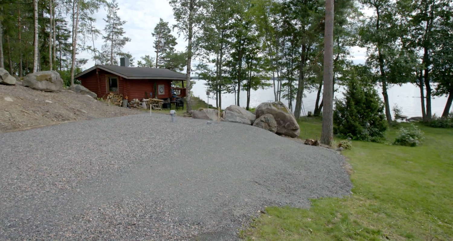 Punainen mökki harmaalla sorapihalla, kivikkoisia rantametsiä ja sininen järvi taustalla