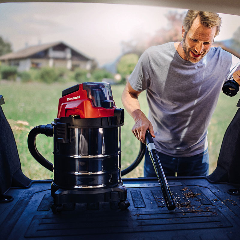 Mies imuroiden puu- ja metsäjätteitä autontavaratilassa Einhell-työimurilla, taustalla niitty ja mökki