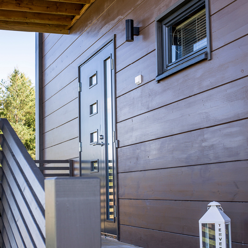 Kuvassa talon seinä, joka on maalattu peittävällä puunsuojalla. Kuvassa myös talon ulko-ovi sekä valkoinen lyhty.