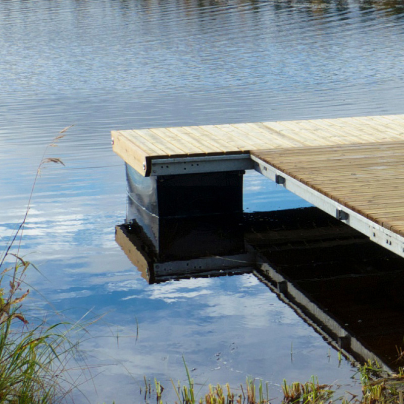 Vaalea puulaituri heijastuu tyynessä sinisessä vedessä, rannassa ruokoja.
