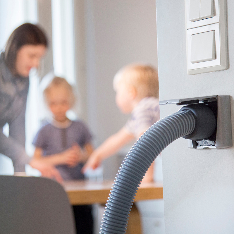 Harmaa keskusimurin letku seinäliitännässä, taustalla äiti ja lapset keittiön pöydän äärellä.