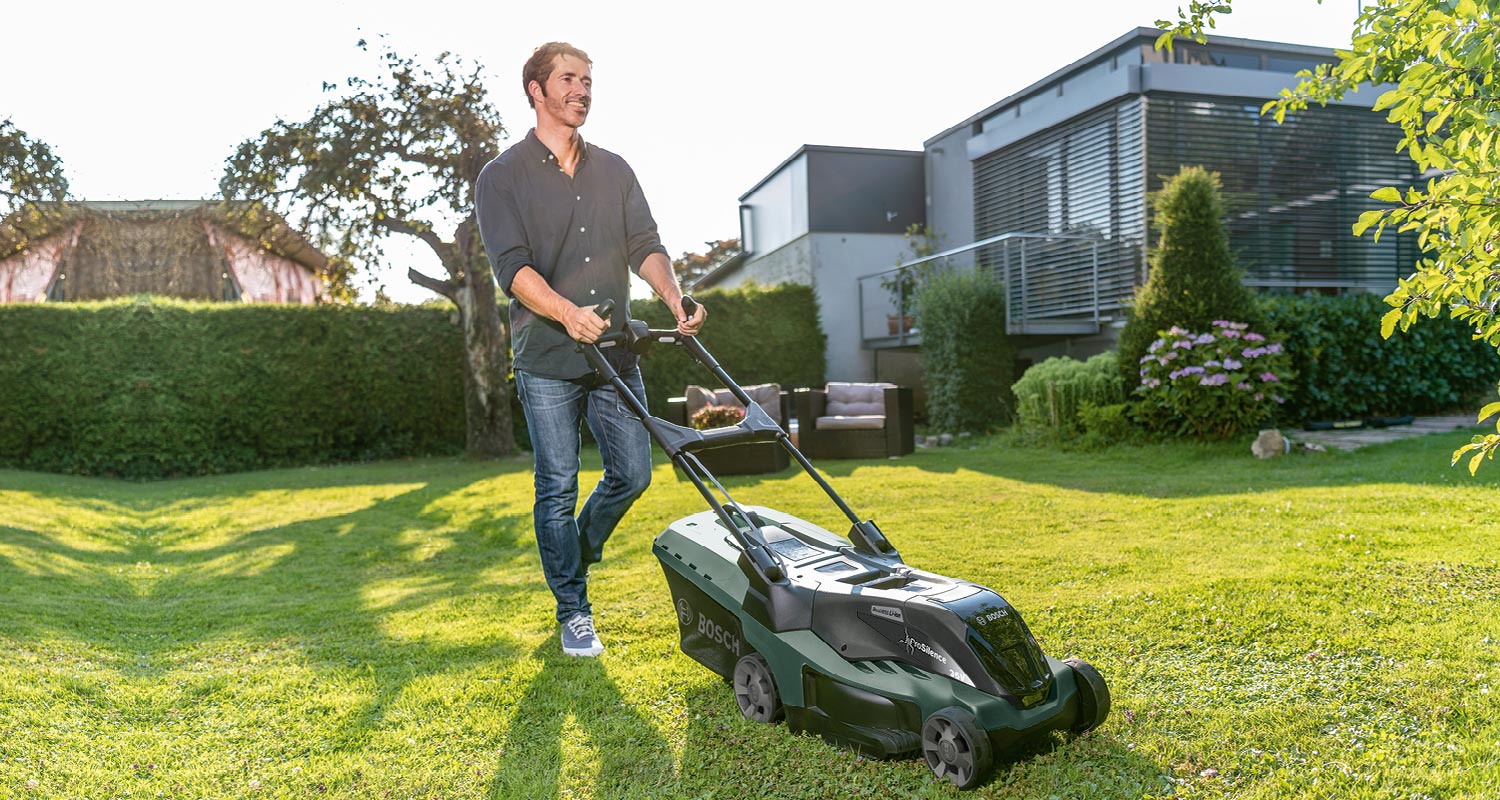 Henkilö työntää vihreää Bosch-ruohonleikkuria aurinkoisella pihalla.