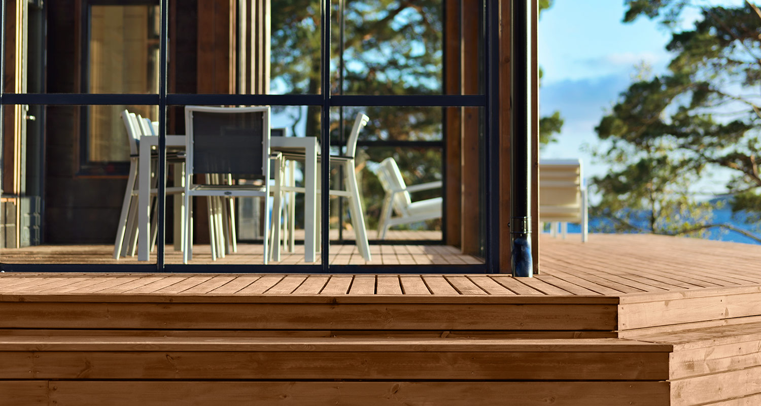 Vaalea puulattia ja lasiseinä, valkoiset terassituolit, metsä- ja järvinäkymä