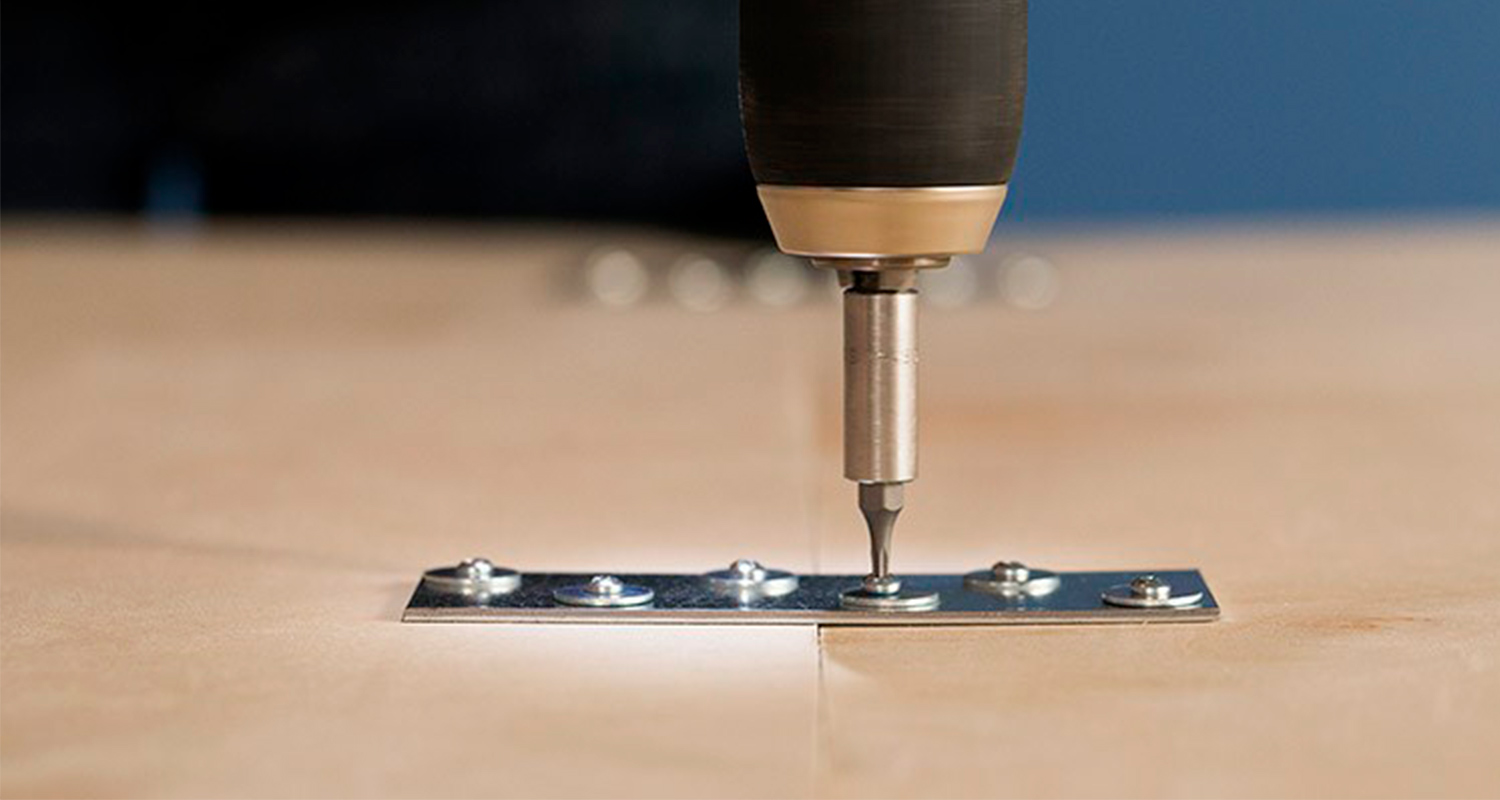Porakoneen metallinen kärki ruuvaa hopeista liitoslevyä kahden vaalean puulevyn saumassa