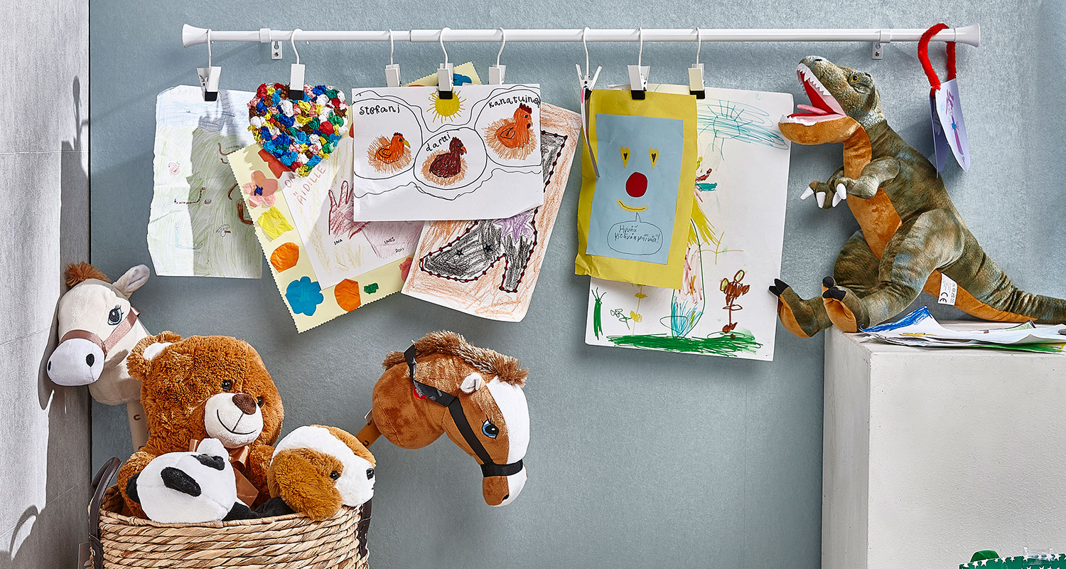 Vaaleansinistä taustaa vasten roikkuu värikkäitä lasten piirustuksia, alla nalle ja dinosaurus.
