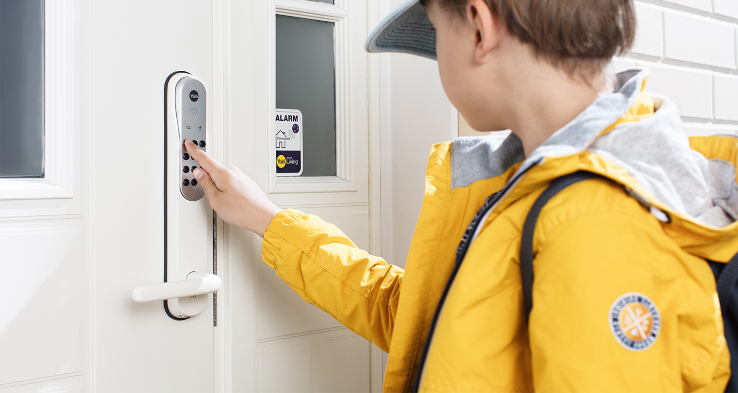 Henkilö keltaisessa takissa näppäilee valkoisen älylukon numeropainikkeita valkoisessa ovessa.