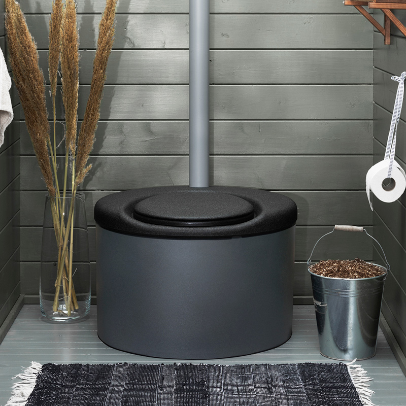 Harmaaseinäisessä kompostivessassa musta istuin, rättimatto, ruokoasetelma ja sanko.