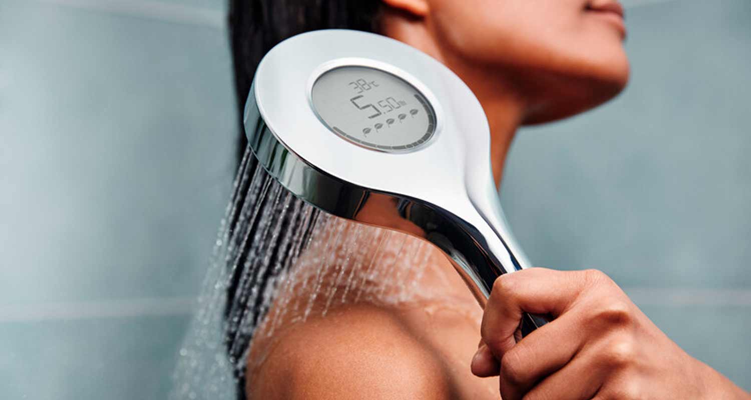 Krominen diginäytöllinen suihkupää suihkuttaa vettä naisen olkapäälle harmaata taustaa vasten.