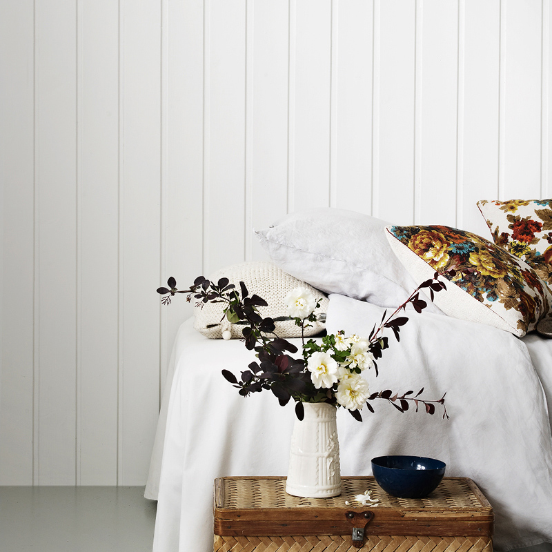 Valkoisessa huoneessa maljakko rottinkipöydällä, valkoiset kukat, viininpunaiset oksat, sininen kulho.