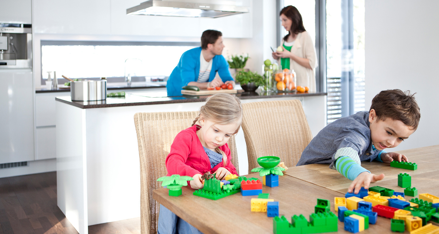 Lapset leikkivät värikkäillä Duplo-palikoilla puupöydän ääressä valkoisessa keittiössä.