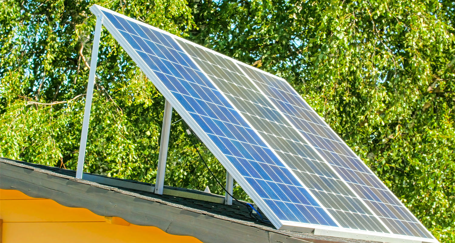 Siniset aurinkopaneelit metallikehikossa keltaisen katon ja vihreiden lehtipuiden edessä.