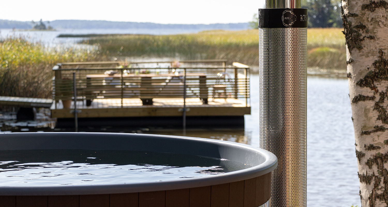 Harmaareunainen puulämmitteinen palju järvenrannalla, metallinen piippu ja puulaituri.