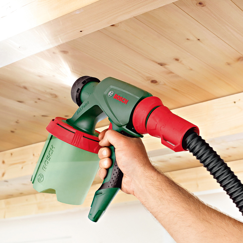 Käsi pitelee vihreä-punaista Bosch-sumutusmaalauskonetta puukattoa maalatessa