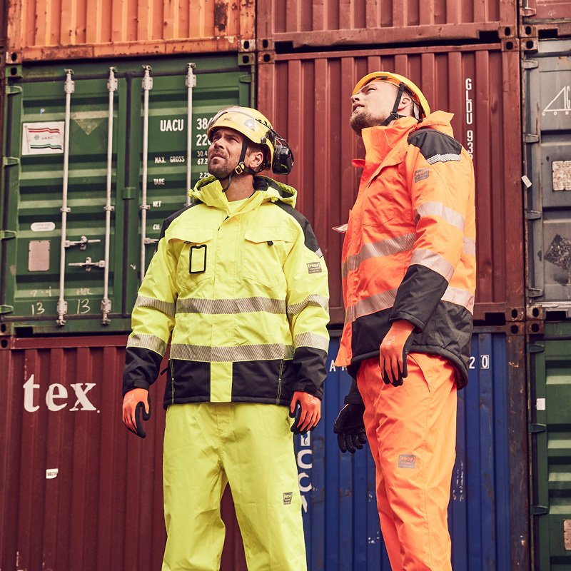 Kaksi satamatyöntekijää neonkeltaisissa ja oransseissa heijastinpuvuissa konttien edessä
