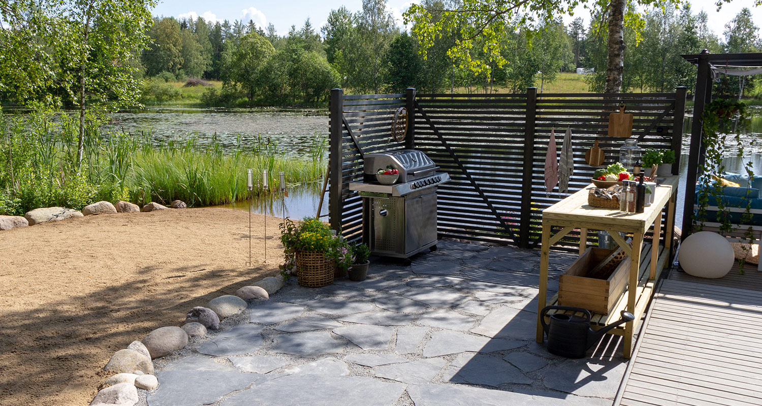 Järvenranta, harmaa kiviterassi, hiiligrilli, puinen työtaso ja musta säleaita, taustalla metsää.