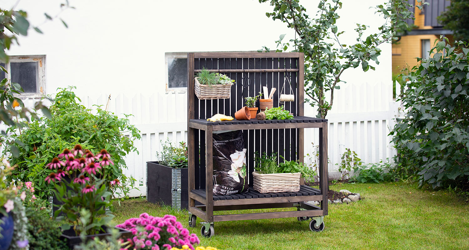 Tumma puinen puutarhatyöpöytä rullilla, ruukkuja ja vihreitä yrttejä valkoisen aidan edessä