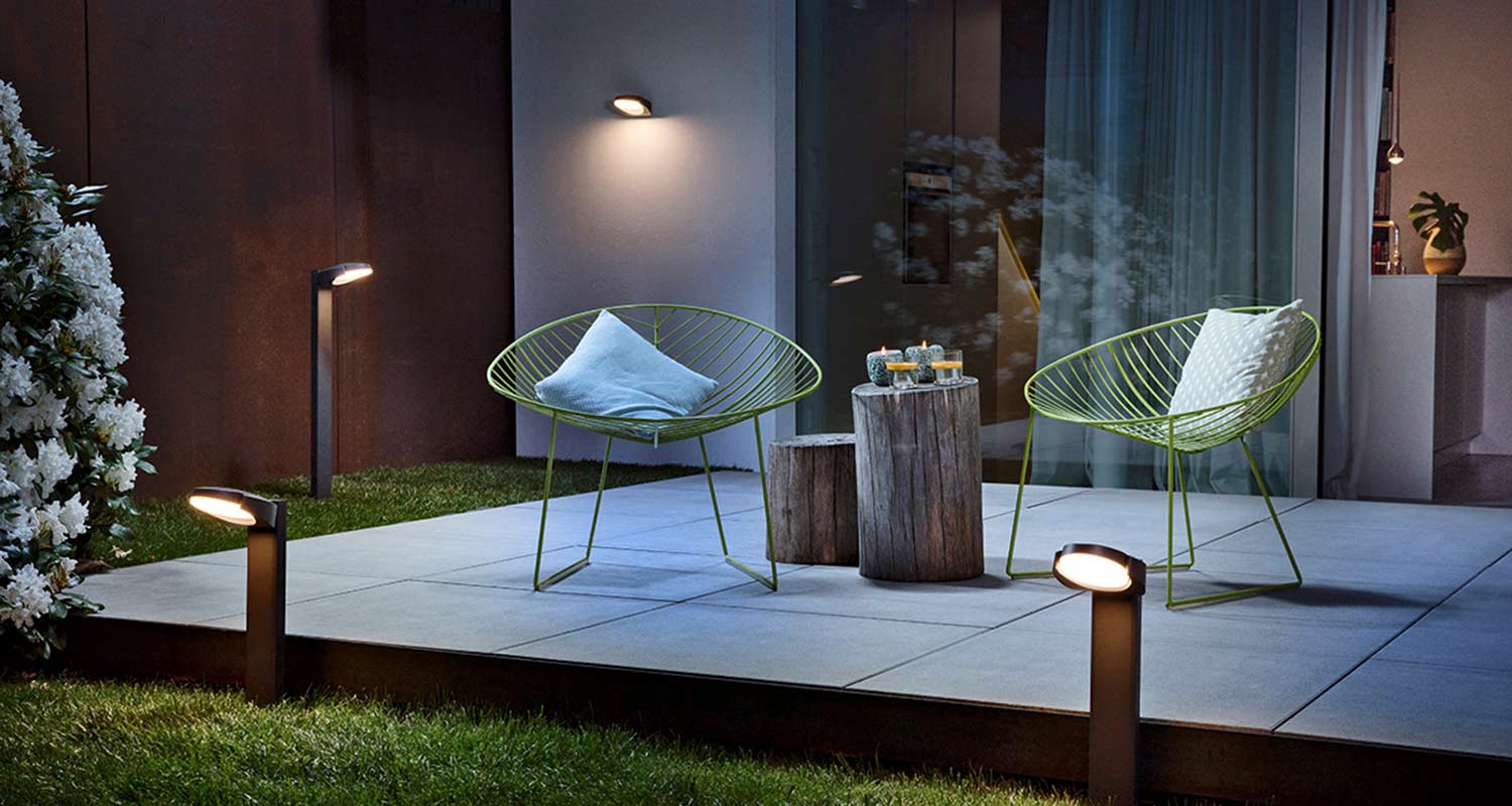 Kahden limenvihreän metallituolin, tyynyjen ja puupölkky-pöydän kynttilöineen valaistu iltaterassi.