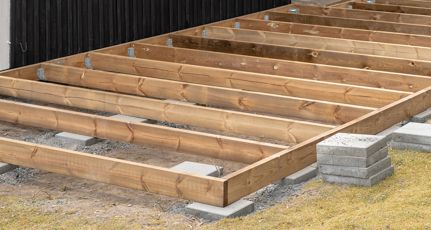 Rakenteilla oleva terassin puukehikko harmailla betonilohkoilla mustan seinän edessä