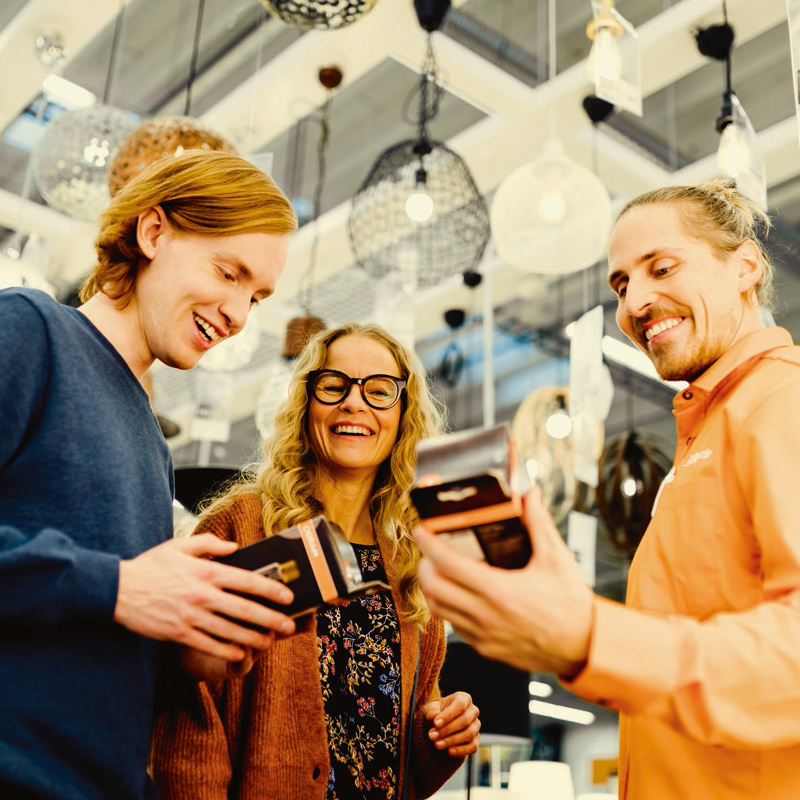 Kolme henkilöä valaistuskaupassa tutkii mustanoransseja lamppupaketteja ja roikkuvia valaisimia