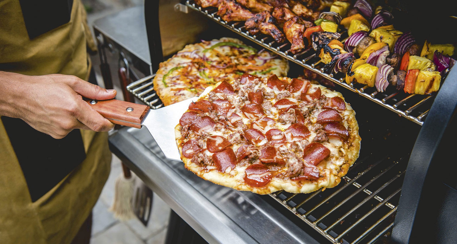 Grillissä paistuva pizza punaisilla pepperonaviipaleilla ja vihannes-lihavartaat, kädessä pizzalapio