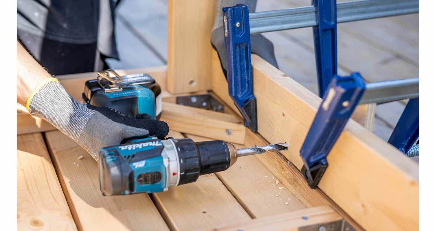 Siniharmaa akkuporakone poraa vaaleaa puulistaa sinisillä puristimilla ja harmaassa käsineessä.