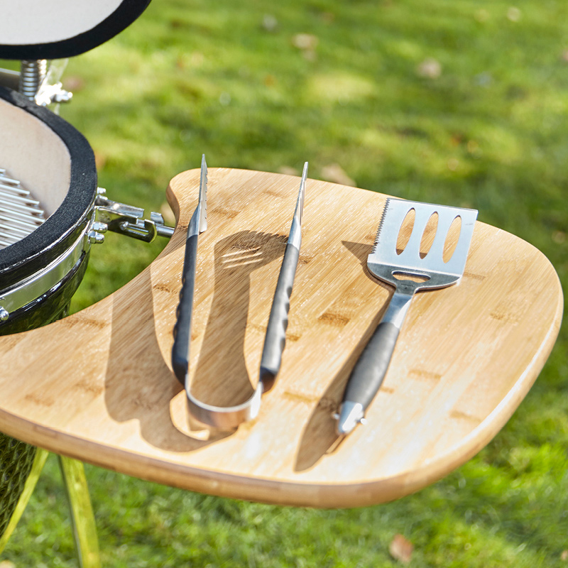 Grillin bambupöydällä metalliset mustakahvaiset pihdit ja lasta, taustalla vihreä nurmikko.