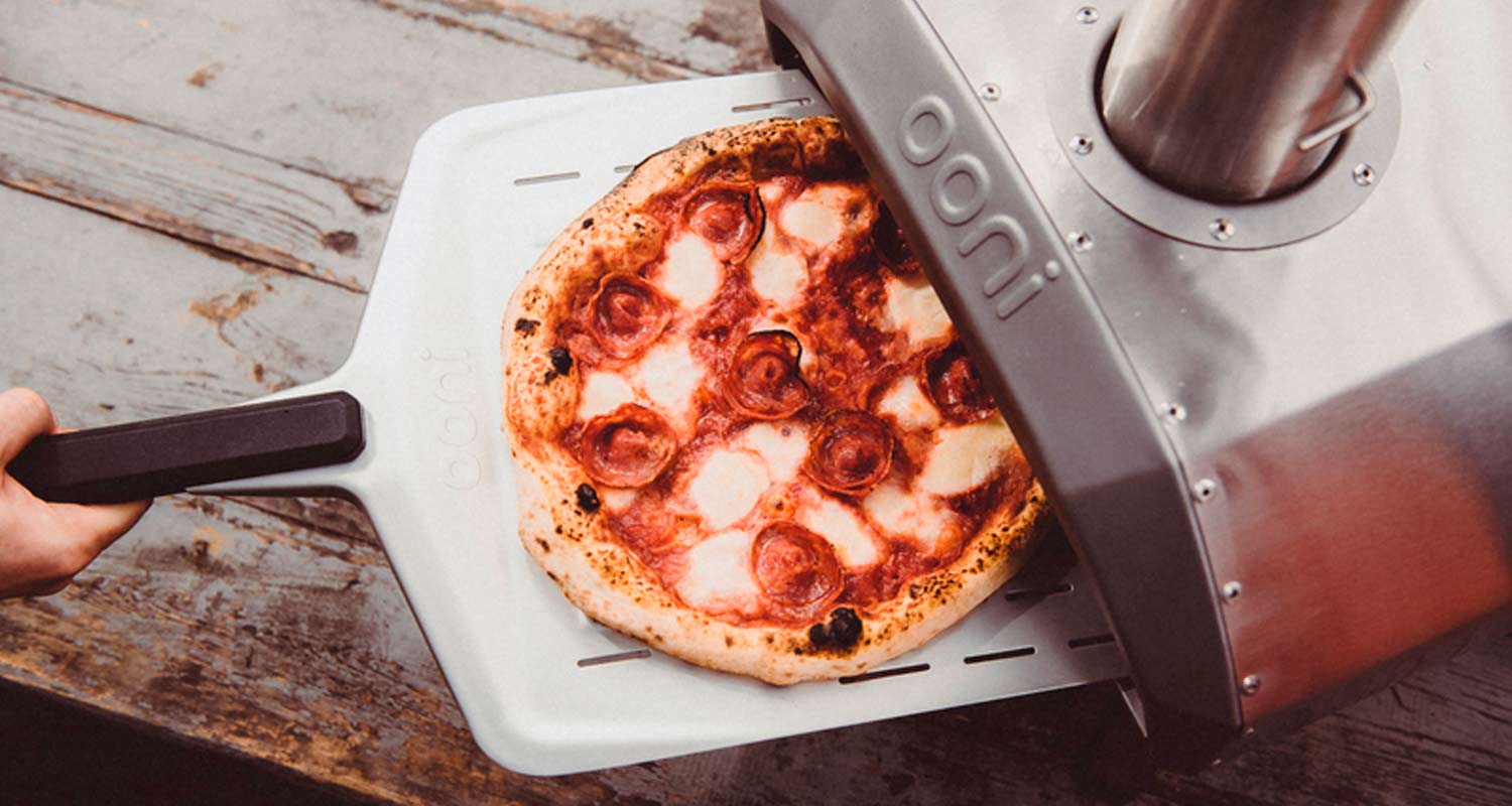 Käsi työntää metallipelillä punavalkoisen mozzarellapepperonipizzan hopeaharmaaseen ulko-uuniin