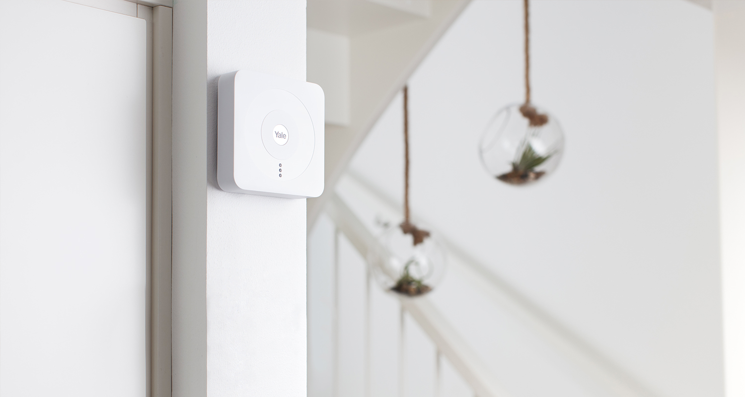 Valkoinen Yale-keskusyksikkö valkoisella seinällä, taustalla portaat ja riippuvat lasiterrariumit.