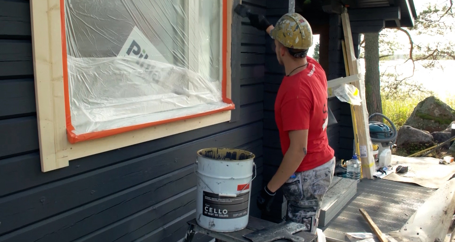 Mies punaisessa paidassa ja kypärässä maalaa tummansinistä puuseinää muovitetun ikkunan vierellä