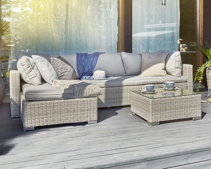 Vaaleanharmaa punottu pihasohva tyynyillä ja pleedillä, edessä lasipöytä kahvikuppeineen.