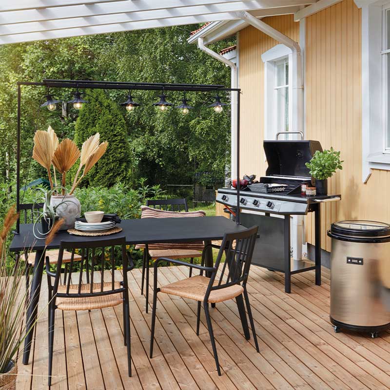 Vaaleankeltaisen puutalon edessä terassi: musta pöytä, punoksiset tuolit, grill ja vihreä puutarha.