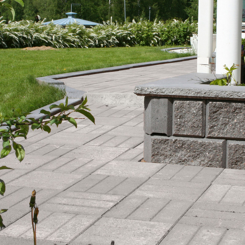 Harmaa betoniterassi tummanharmaalla reunakivellä, taustalla nurmikko ja valkoiset kukkapuskat