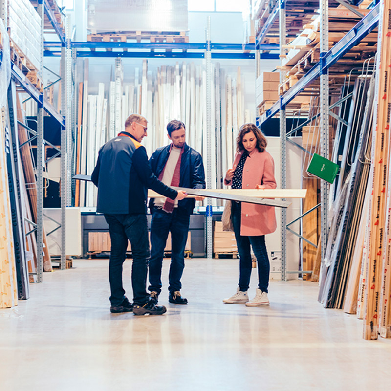 Rautakaupan hyllyjen välissä myyjä ojentaa vaaleita puulautoja kahdelle asiakkaalle.