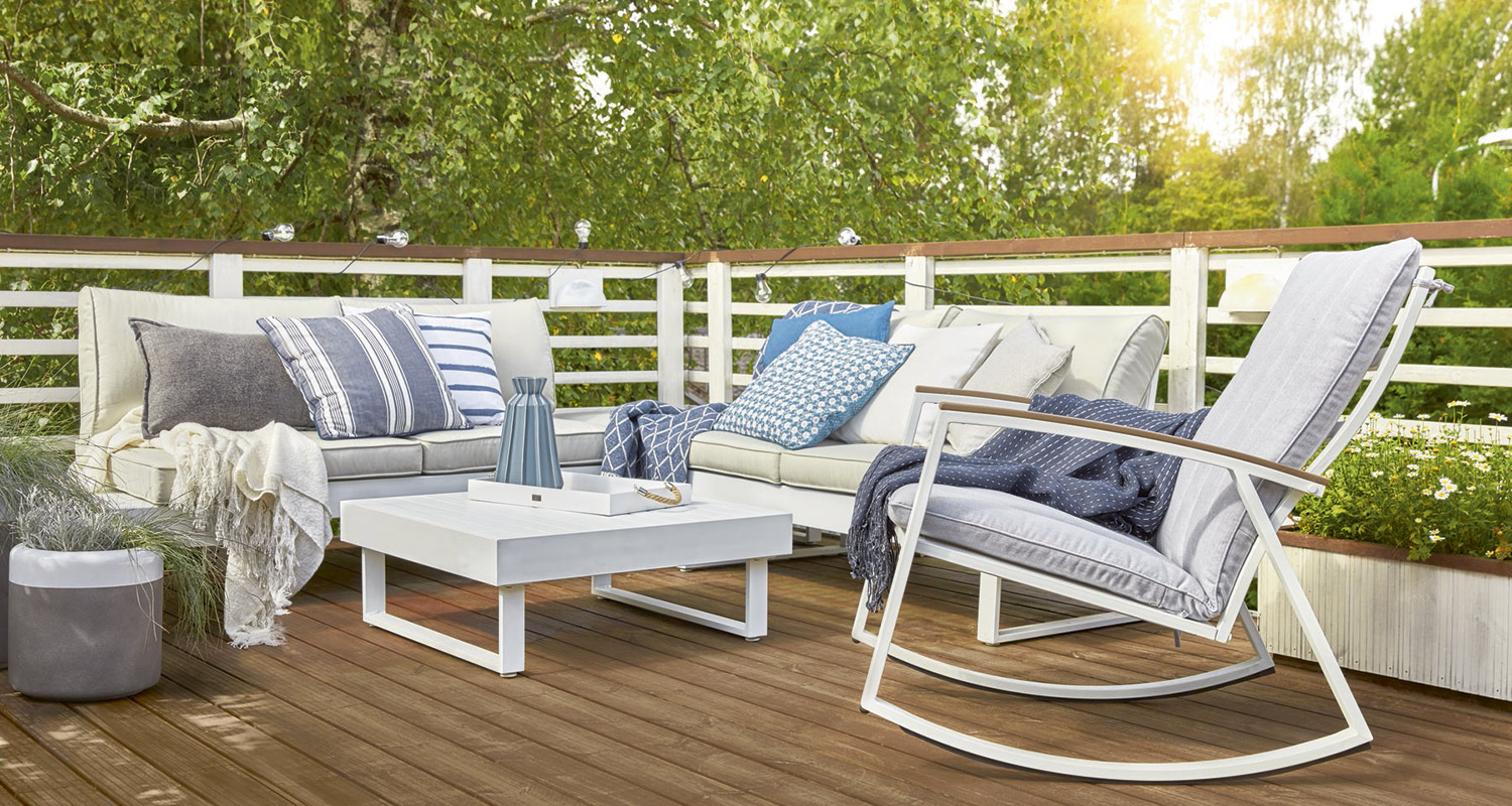 Aurinkoinen puulattiaterrassi, valkoinen kulmasohva sinivalkoisin tyynyin ja harmaa keinutuoli.