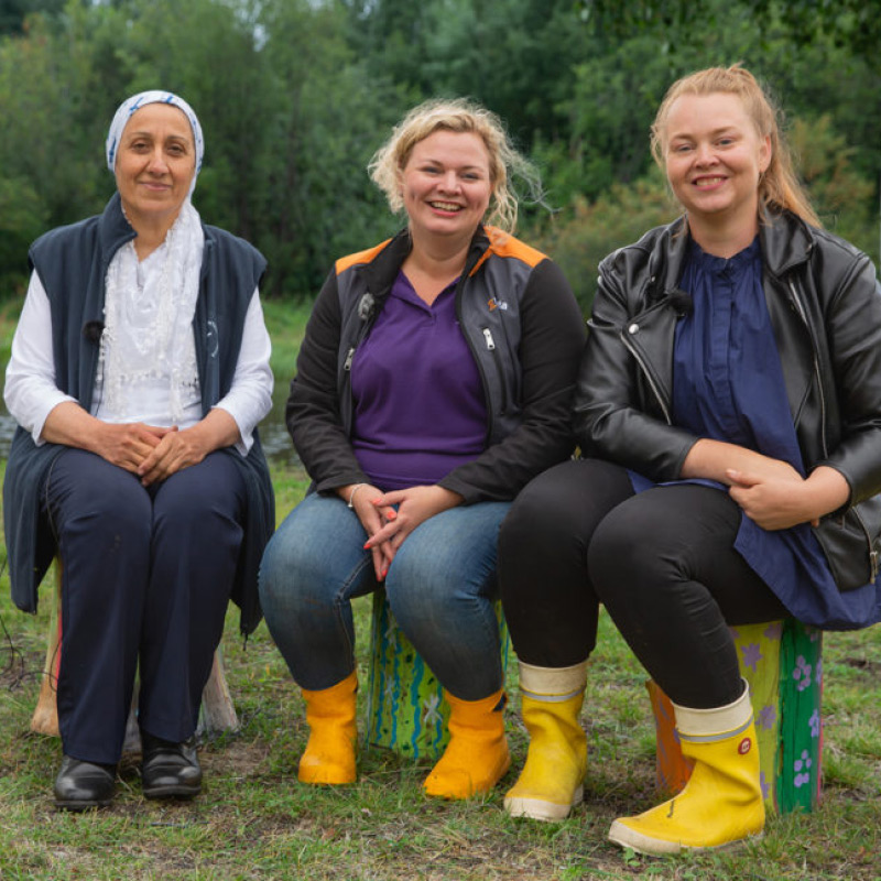 Kolme naista istuu maalatuilla penkeillä niityllä, kaksi keltaisissa kumisaappaissa.