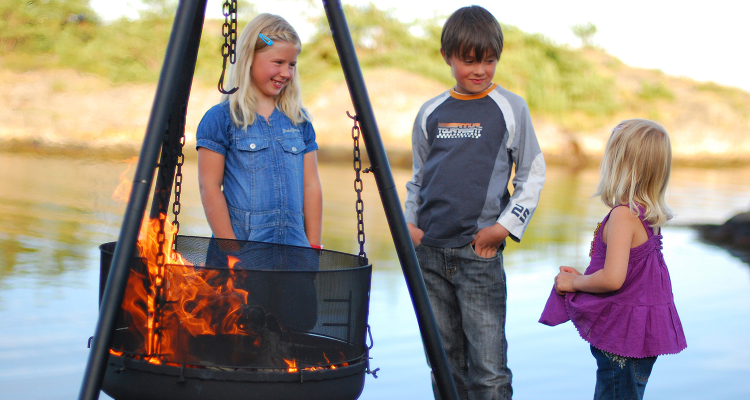 Kolme lasta järven rannalla katsovat tulen leimuavaa kolmijalkaista nuotiokeitintä