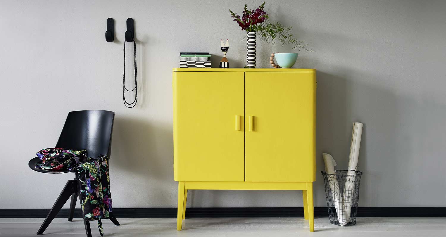 Keltainen lipasto harmaata seinää vasten, musta tuoli kukkahuivilla, koristeita ja rullia korissa.