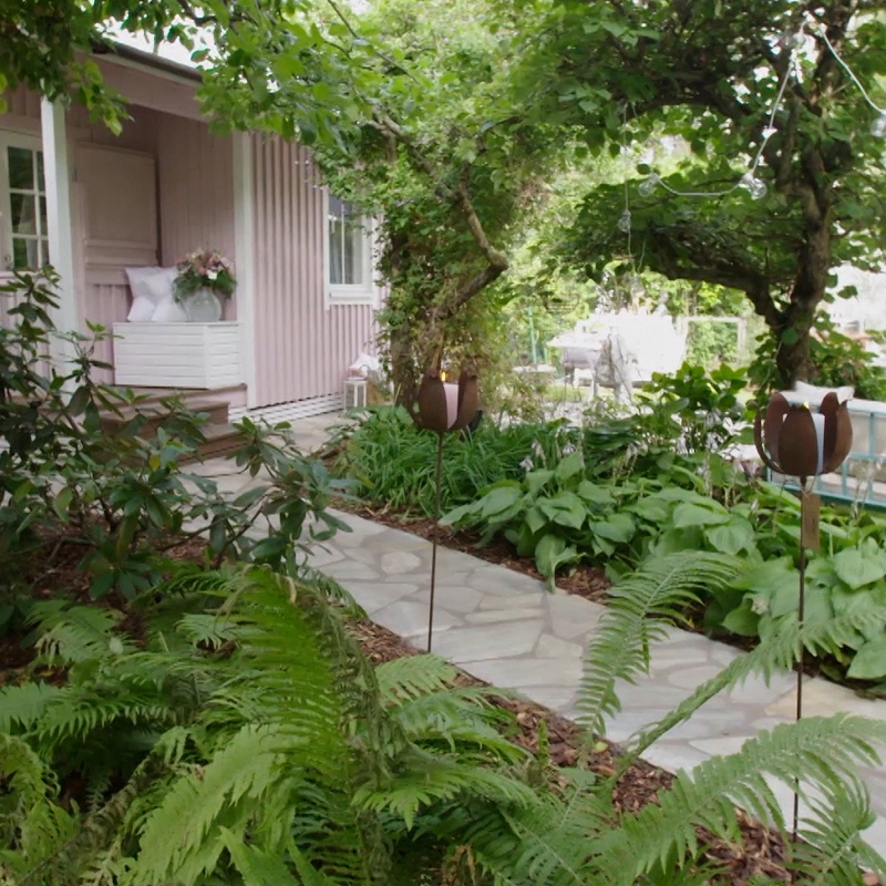 Kivipolku kaartuu rehevän vihreän puutarhan halki kohti vaaleanpunaista mökkiä.