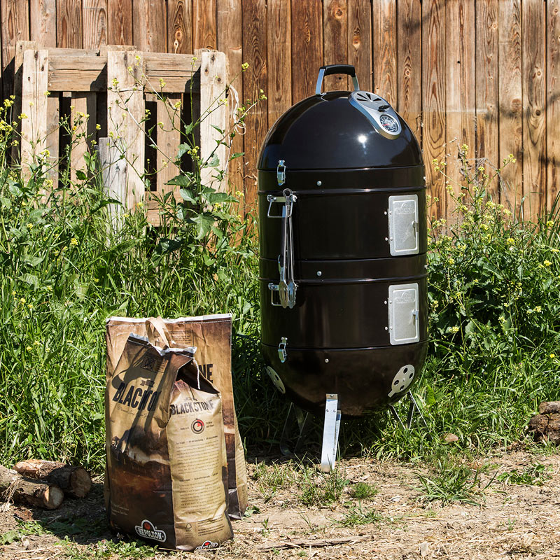 Musta pystygrilli-savustin ja ruskeat hiilipussit nurmikolla puuaitaa vasten