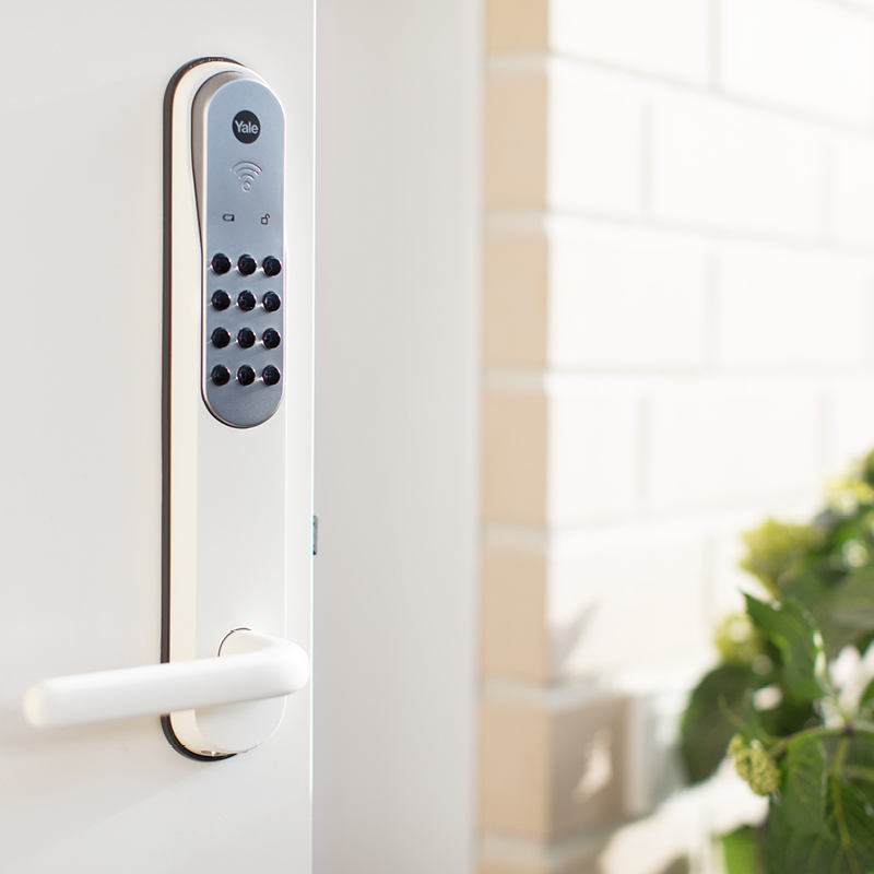 Valkoiseen oveen asennettu Yale-sähkölukko ja -kahva, taustalla vaalea tiiliseinä ja viherkasvi