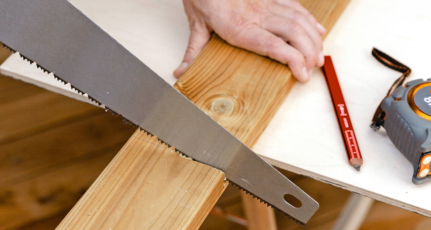 Vaaleaa puulistaa sahataan käsisahalla, vieressä punainen merkintäkynä ja mittanauha.