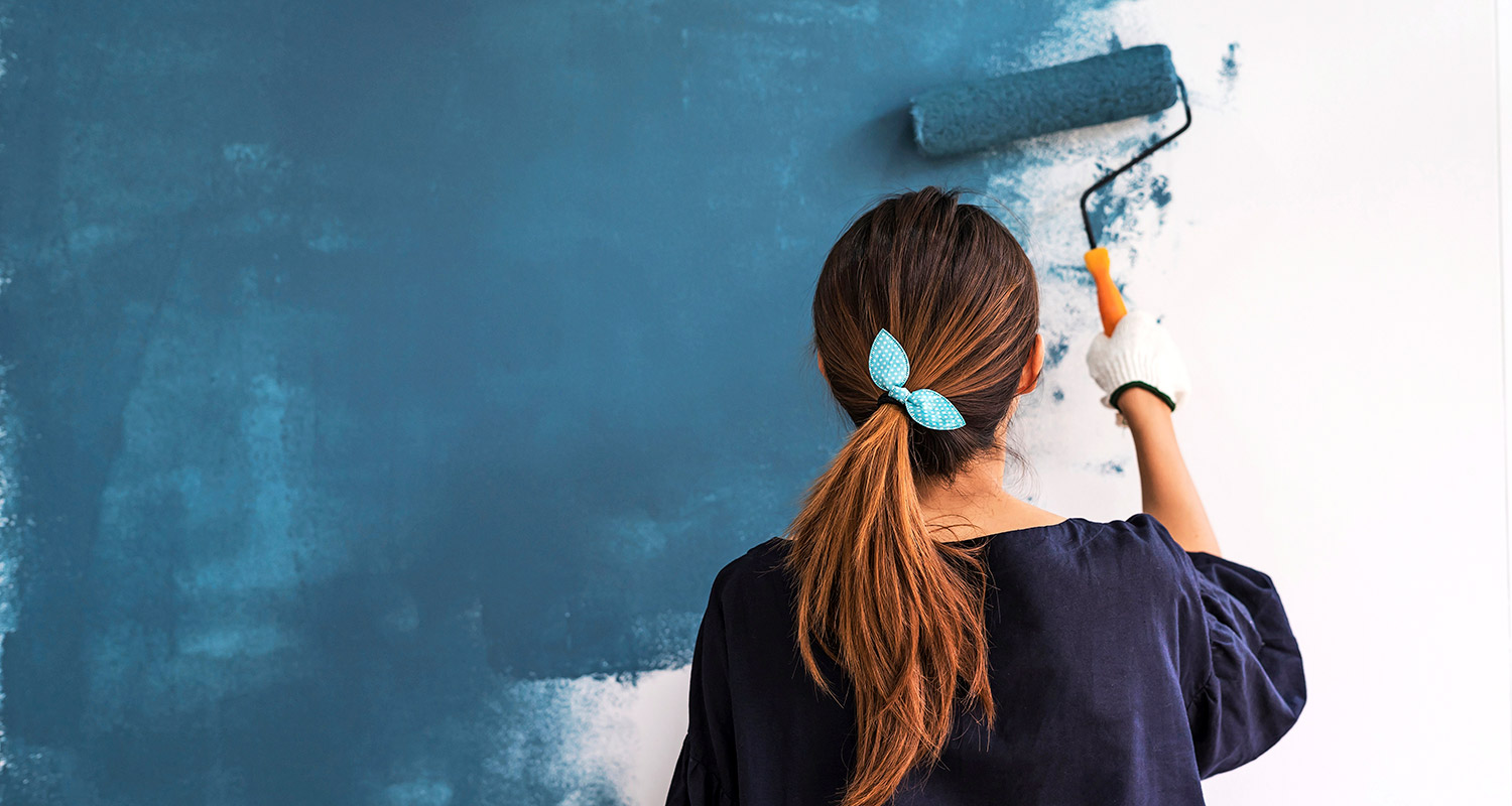 Nainen selin maalaa valkoista seinää tummansinisellä maalirullalla, turkoosipannalla.