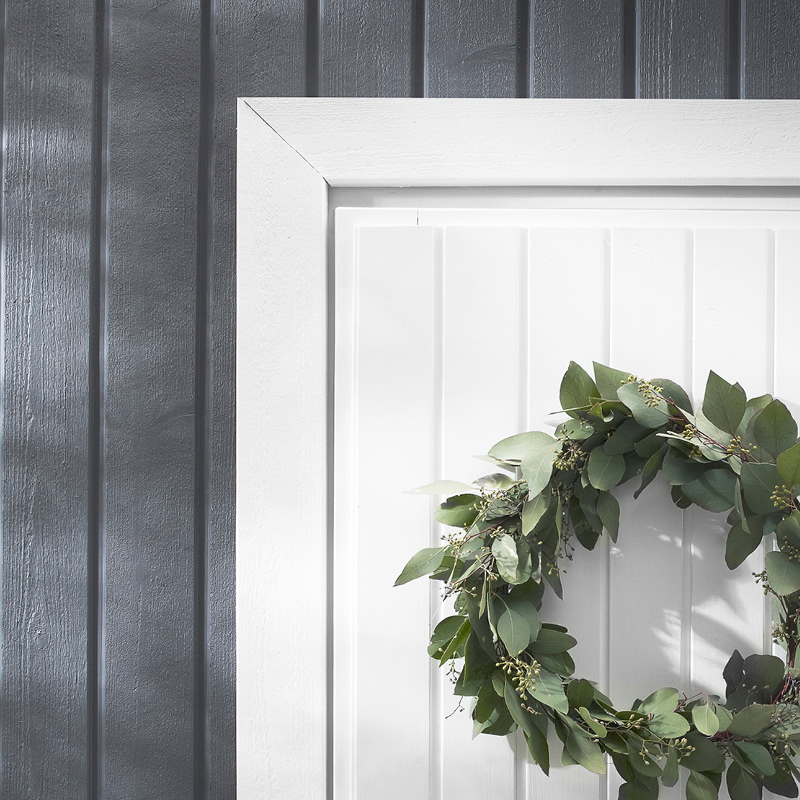 Kuvassa on harmaata talon ulkoseinää sekä osa ovea joka on maalattu valkoiseksi kalustemaalilla. Ulko-ovessa on lehdistä tehty kranssi.