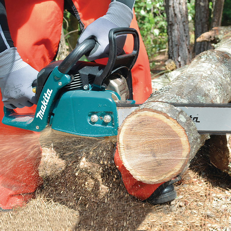 Henkilö punaisissa suojahousuissa sahaa puupölkkyä turkoosilla moottorisahalla metsässä.