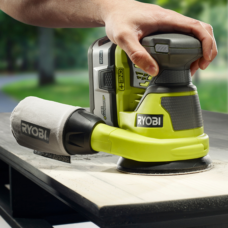 Käsi pitelee vihreää Ryobi One+ akkukäyttöistä hiomakonetta puupinnan päällä.