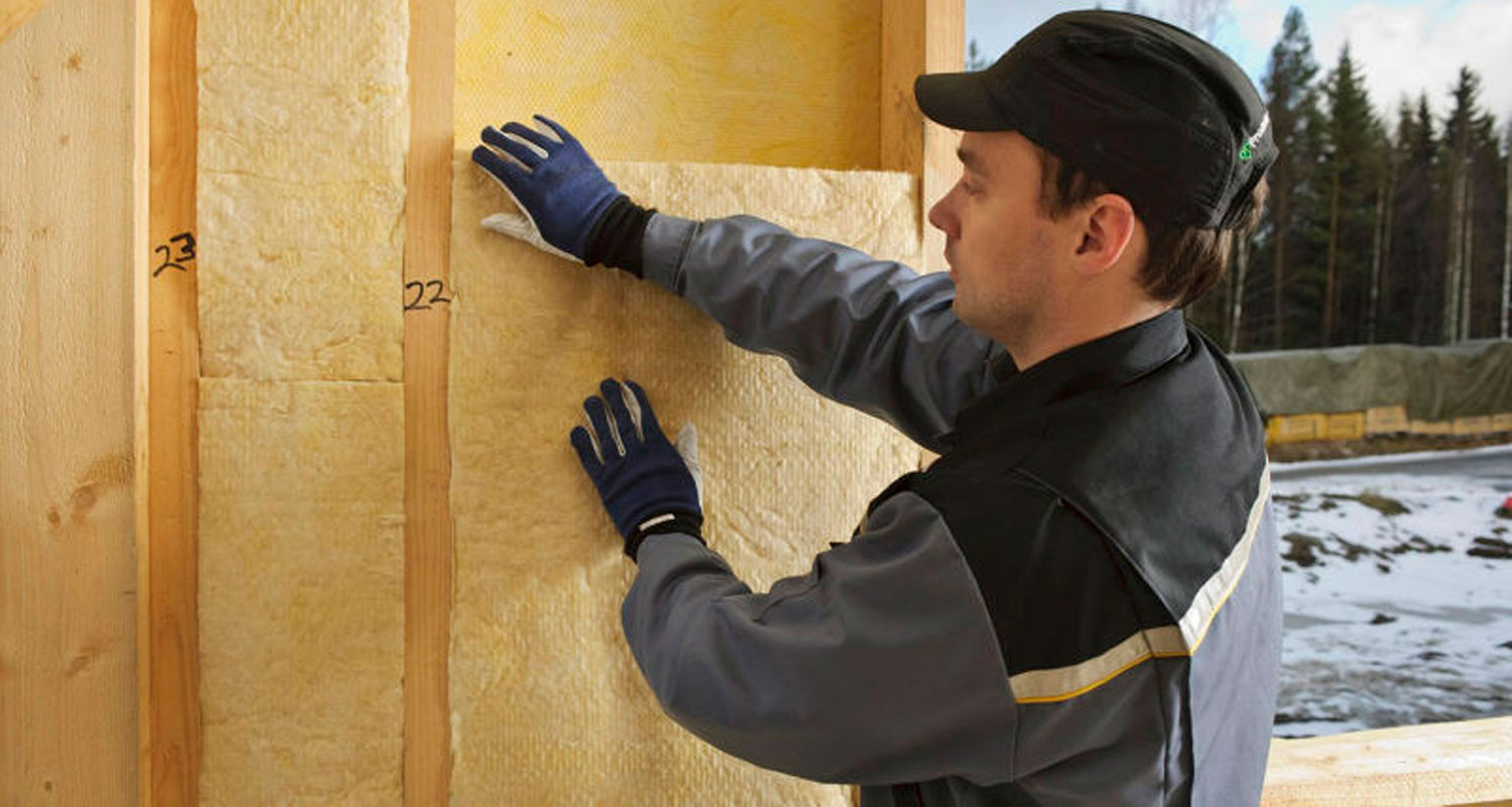 Työmies kiinnittää keltaista lämmöneristettä puurunkoon lumisessa rakennusympäristössä.